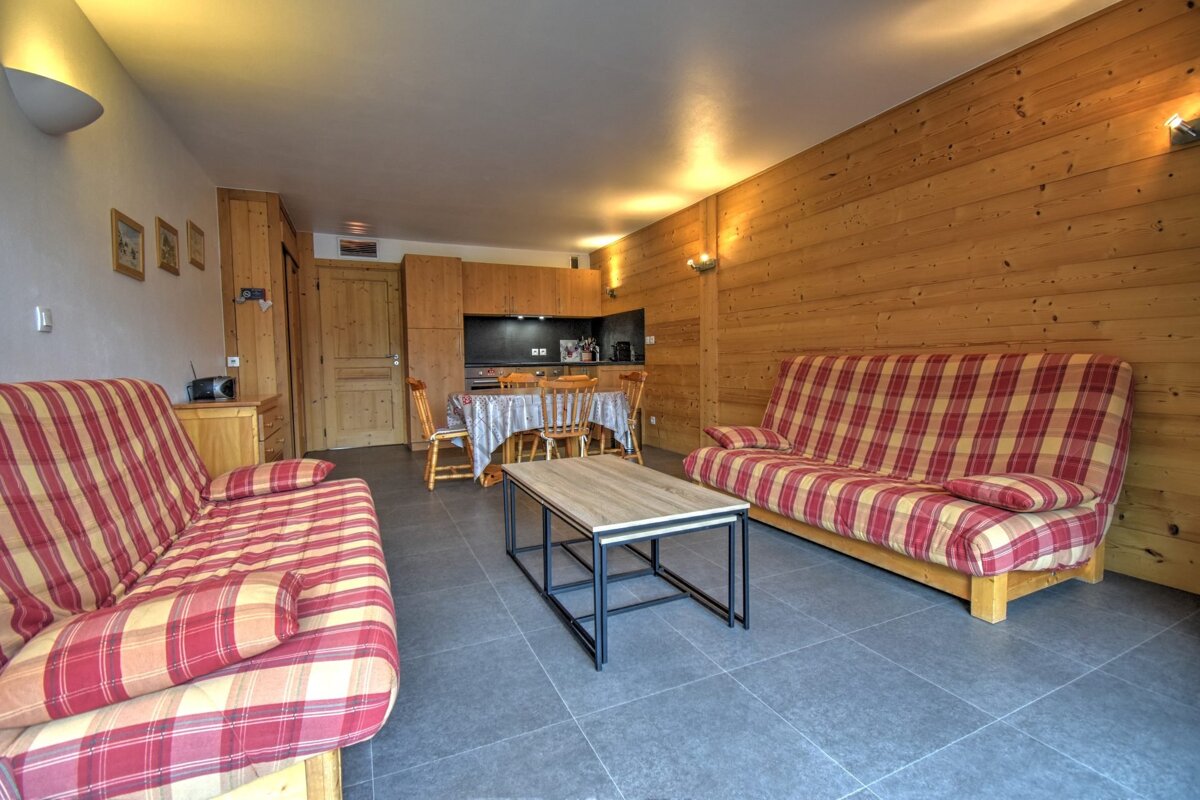 An open-plan living room/kitchenette with two red and yellow plaid sofas, wooden walls, a dining table, and a dark tiled floor.