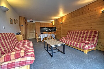 An open-plan living room/kitchenette with two red and yellow plaid sofas, wooden walls, a dining table, and a dark tiled floor.