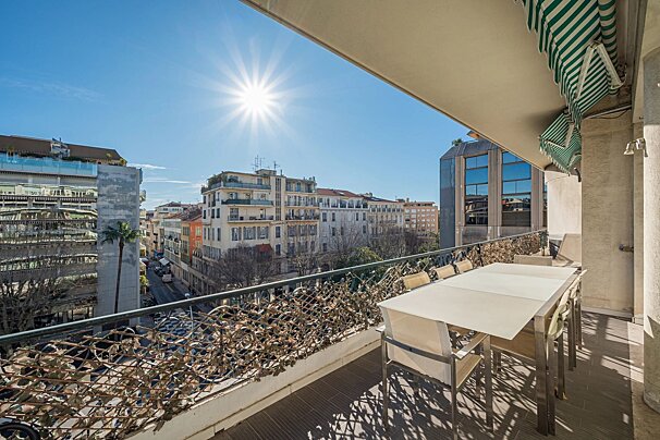 A sunny balcony with an outdoor dining table overlooks a vibrant city street lined with buildings under a clear blue sky.