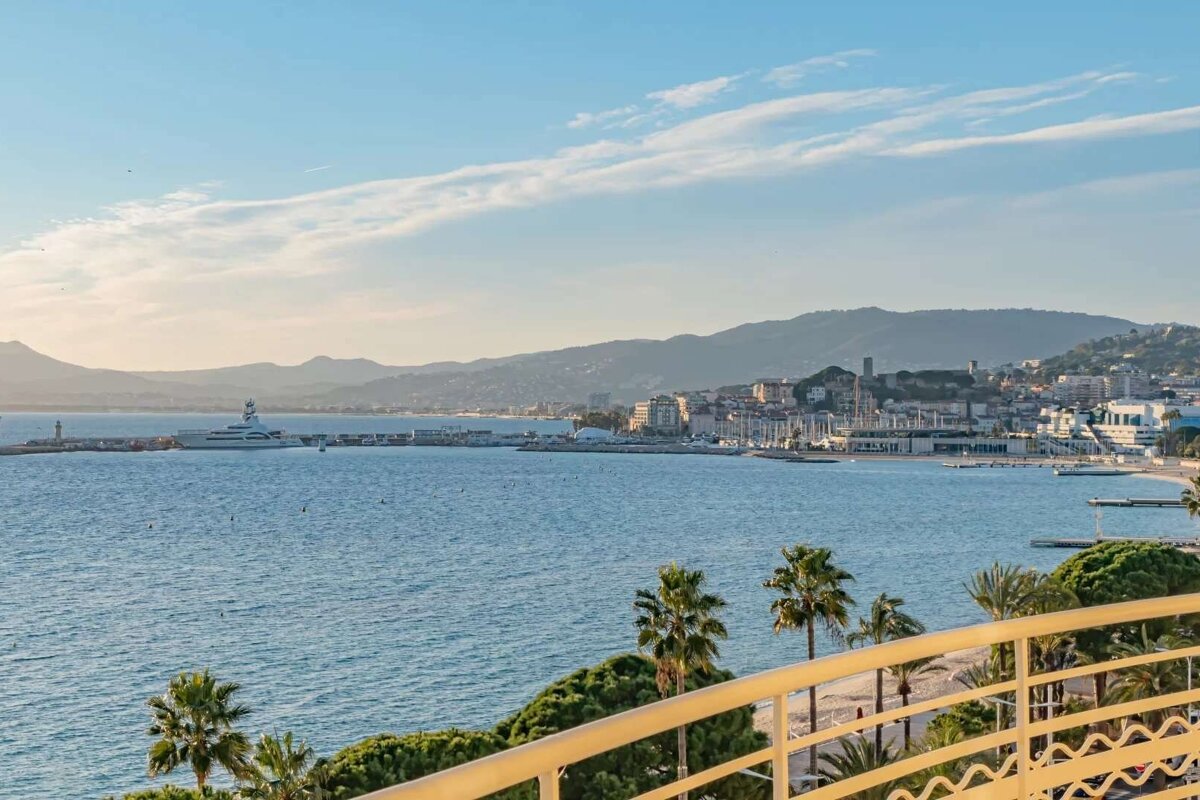 Panoramic view of a sunny coastal city with blue sea, mountains, and a large yacht. Palm trees and a balcony frame the bustling marina.