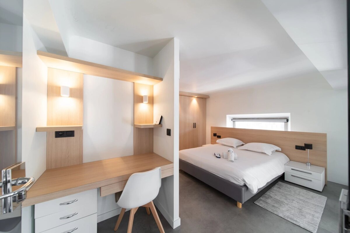 A modern minimalist room featuring a light wood desk with a white chair next to a bed with white bedding and wooden headboard.