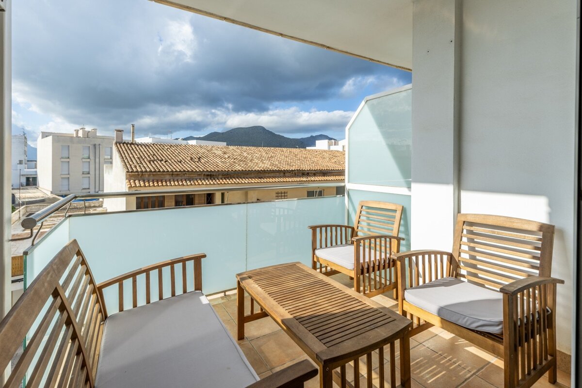 A sunny balcony with wooden seating and a table, overlooking tiled roofs, white buildings, and distant mountains under a cloudy sky.