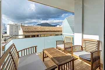 A sunny balcony with wooden seating and a table, overlooking tiled roofs, white buildings, and distant mountains under a cloudy sky.