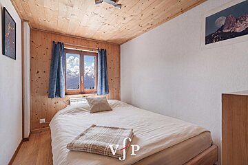 A cozy alpine bedroom featuring wood-paneled walls, a wooden ceiling, and a window framing snowy mountain peaks.