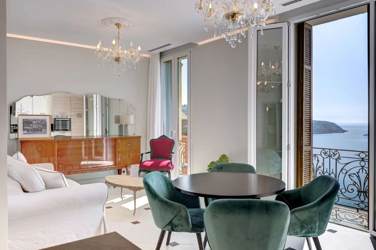 Elegant living/dining room with chandeliers, green velvet chairs, and a balcony overlooking a beautiful sea and distant island.