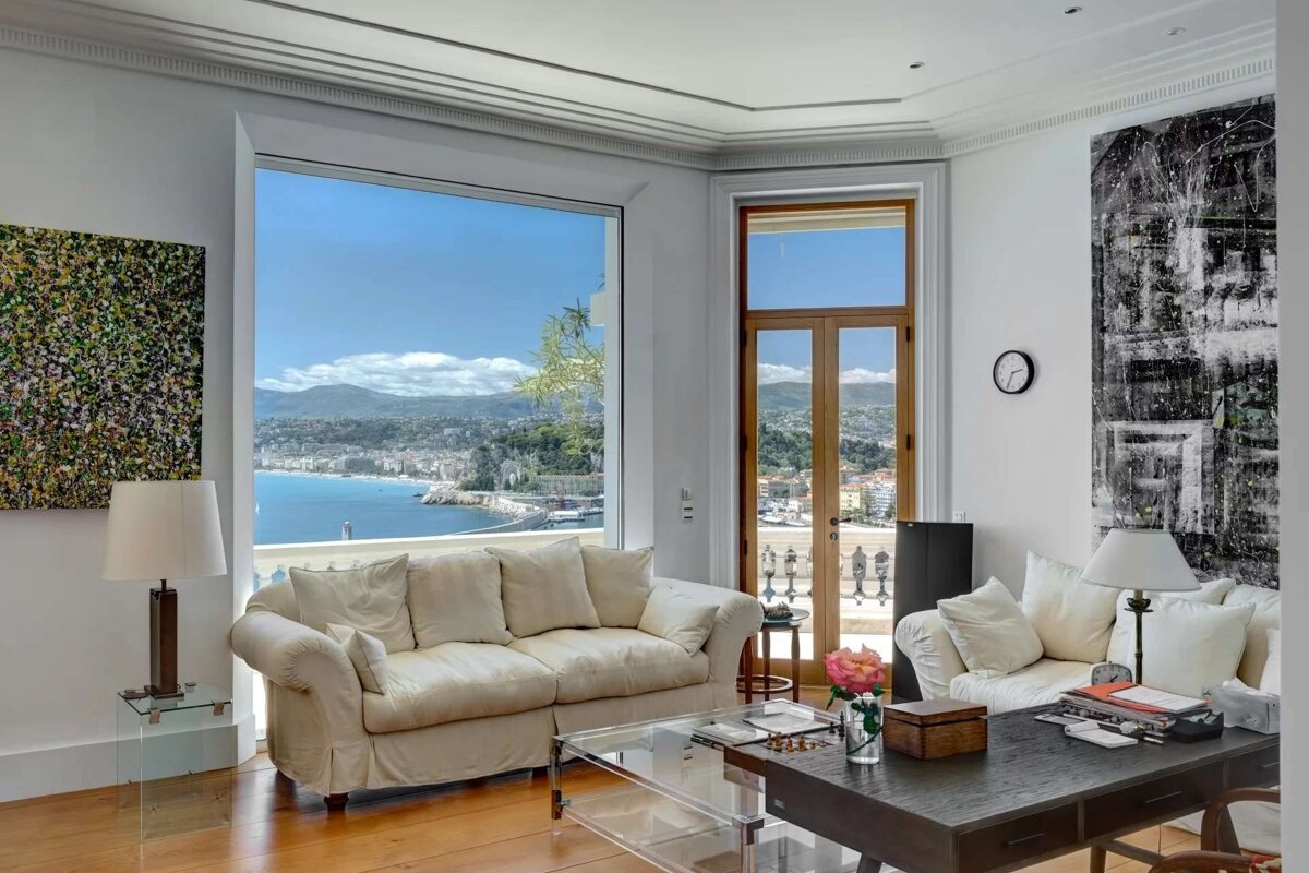 A bright living room with large windows showcasing a stunning panoramic view of a coastal city, mountains, and the blue sea.