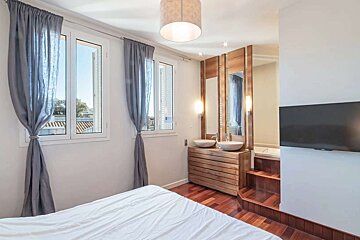 A modern bedroom features a white bed, two windows, and an open-plan bathroom with dual sinks, mirrors, a tub, and a wall-mounted TV.