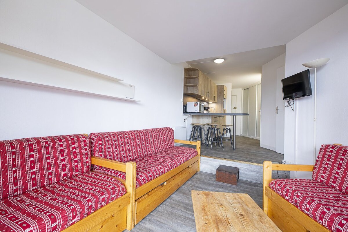 A studio apartment's living room with red patterned sofas, a wooden table, and an elevated kitchenette in the background with a TV.