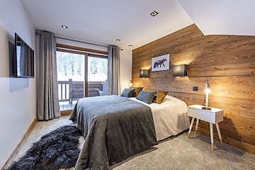 Modern chalet bedroom with a rustic wood accent wall, grey bed, and a balcony overlooking a snowy landscape. Includes a TV and fur rug.