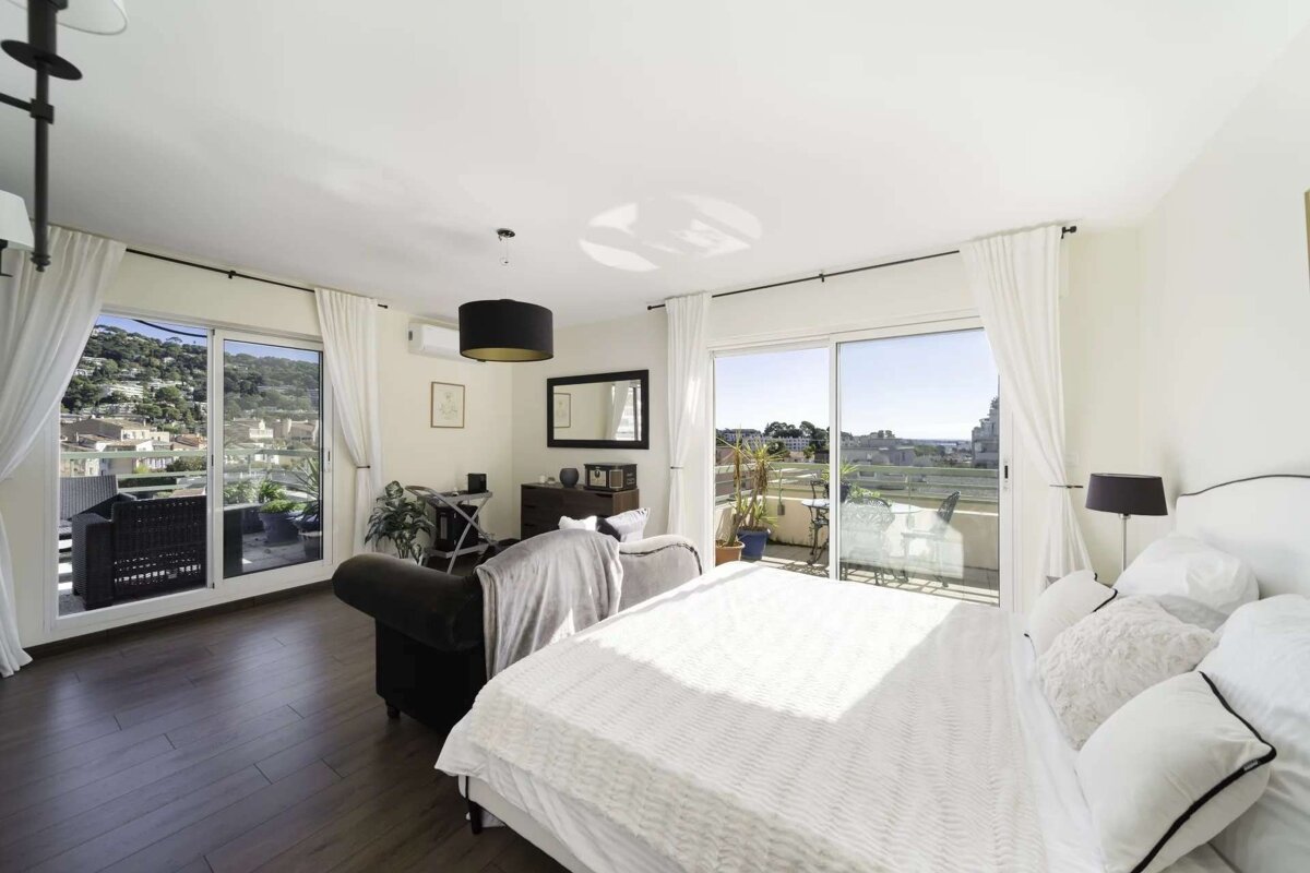 Bright studio room with a bed, living area, and large glass doors opening to a balcony with scenic views of hills and a town.