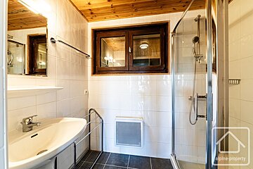 A cozy bathroom with white tiled walls, a wooden ceiling, a sink, mirror, shower cabin, and a rustic wooden-framed window.