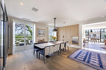 A modern dining room featuring a long table with patterned chairs, wooden floors, large doors, and a blue patterned rug.