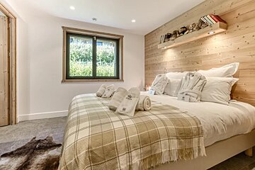 A cozy bedroom featuring a wooden accent wall, a plaid blanket, folded towels, and a window looking out to greenery.