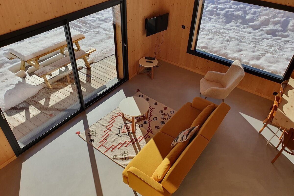 Cozy, sunlit cabin living room with a yellow sofa, patterned rug, and large windows looking out at a snowy deck.