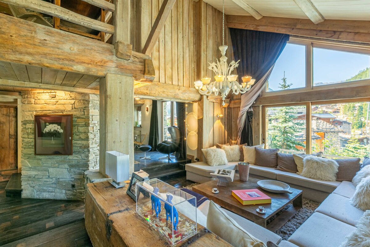 Cozy, rustic living room in a luxury chalet featuring exposed wood beams, a stone wall, comfortable sofa, and large scenic windows.