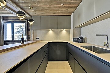 A modern U-shaped kitchen with dark base cabinets, light uppers, wooden ceiling, and a window with snowy trees. Champagne on the counter.