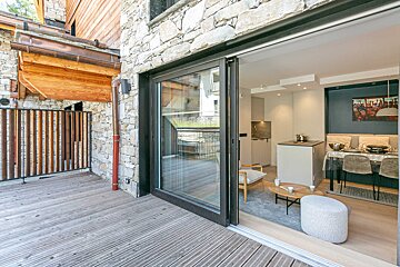 Outdoor wooden deck and stone wall beside a sliding glass door, revealing a modern living/dining area with a kitchenette.