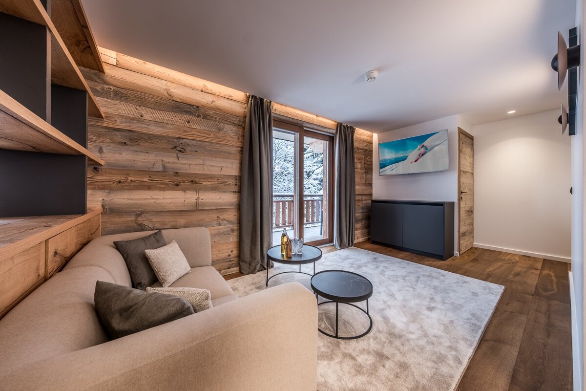 A modern chalet living room with wood paneling, a beige sectional sofa, TV, and a balcony view of a snowy landscape.