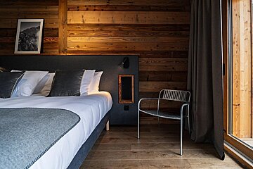 A cozy, modern bedroom with wooden walls/floor, featuring a bed with gray/white linens, mountain artwork, a minimalist chair, and a patio door.