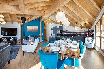 Modern chalet interior featuring a dining table with vibrant blue chairs under a beamed ceiling, alongside a cozy living area.