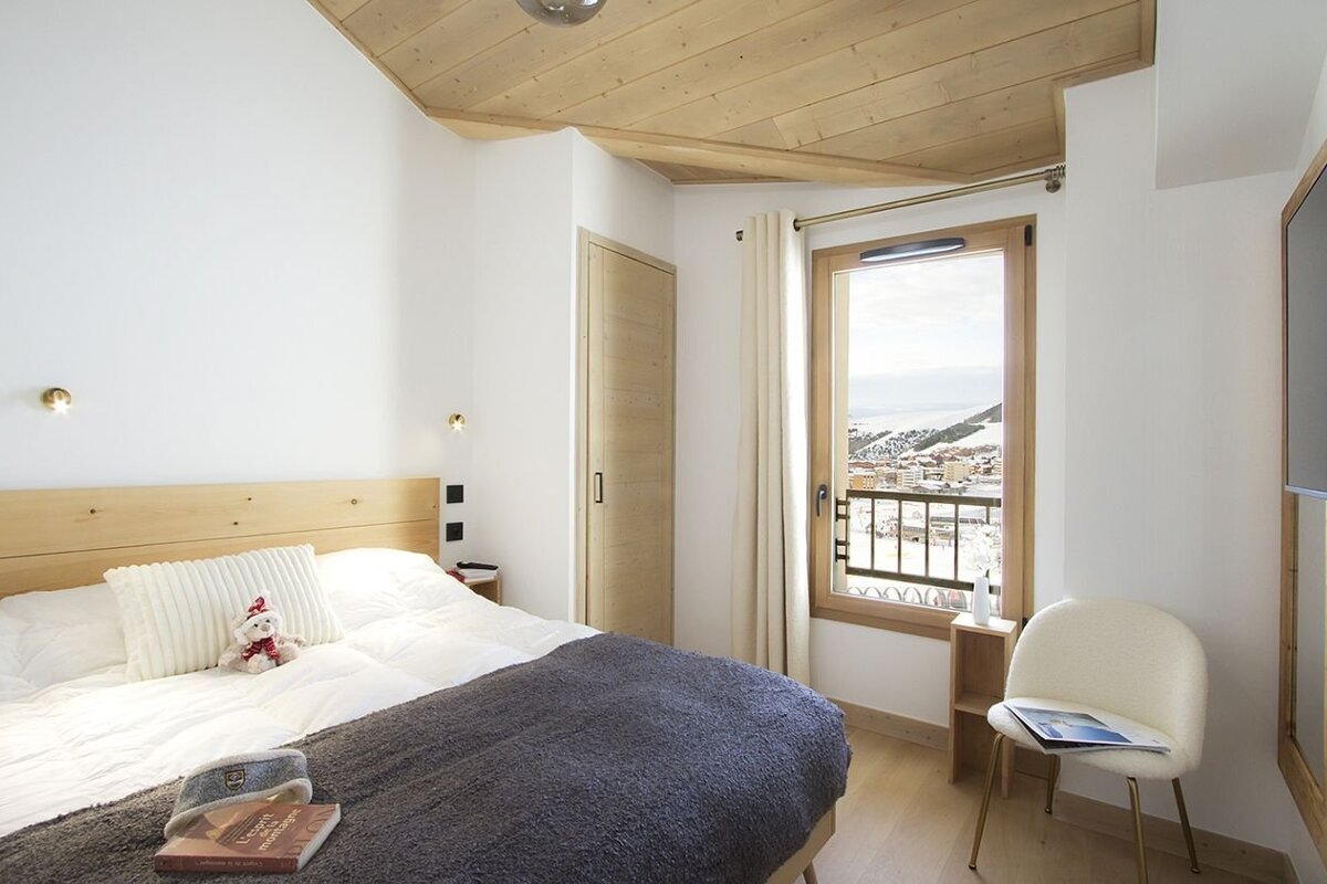 A cozy, modern bedroom with a wooden ceiling, white walls, and a bed, offering a stunning view of a snowy mountain village from its window.