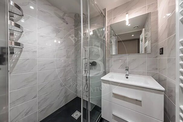 A bright, modern bathroom featuring white marble-effect tiles. It includes a glass-enclosed shower with a dark base, a white vanity, sink, and mirror.