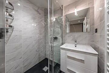 Modern bathroom with white marble-look tiles, a glass shower cabin with a dark base, and a white vanity with a sink and mirror.