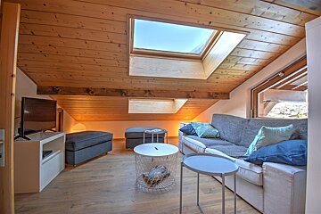 A modern, cozy attic living room with a sloped wooden ceiling, large skylight, gray sofa with blue pillows, and contemporary tables.