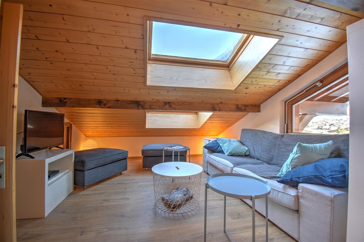 Cozy attic living room featuring slanted wood ceilings with skylights, a grey sofa, ottomans, and small round tables.