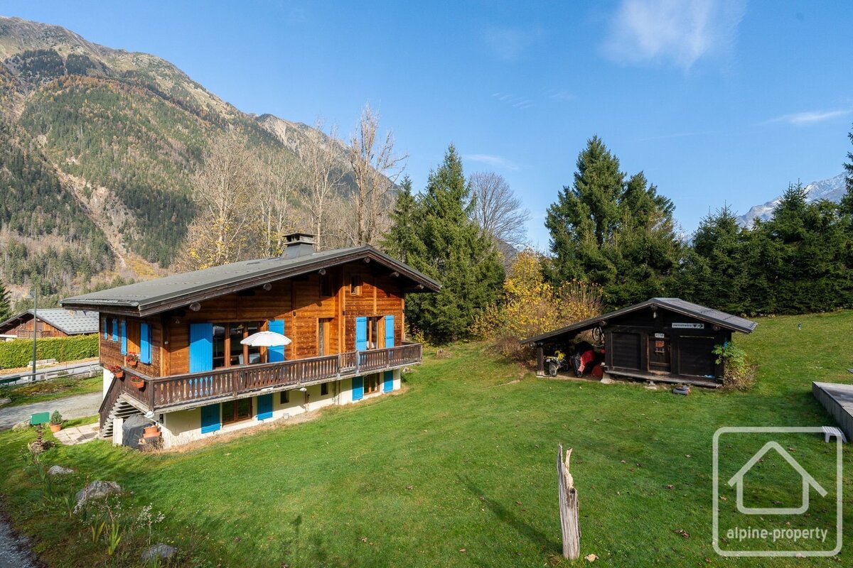 A wooden chalet with blue shutters and a small shed in a grassy yard, surrounded by pine trees and majestic mountains under a blue sky.