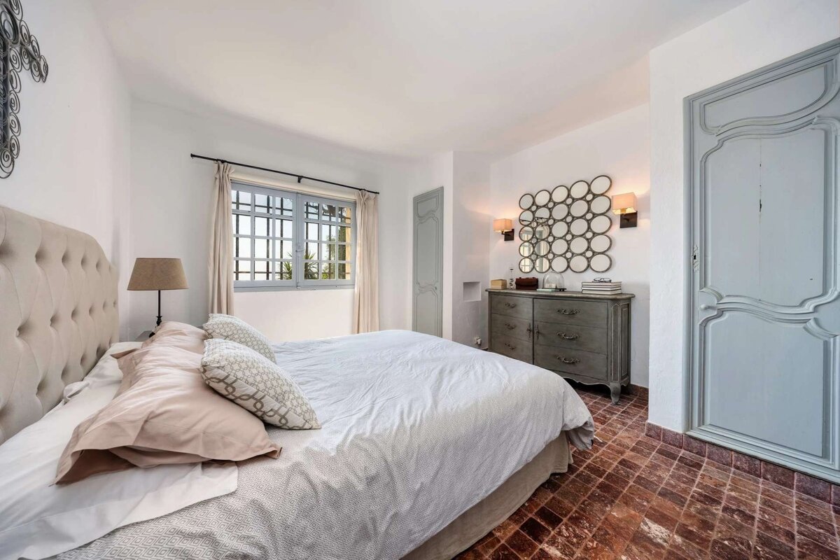 Bright, elegant bedroom with a tufted bed, grey-framed window, antique dresser, unique circle mirror art, and warm terracotta floor. Grey paneled door.