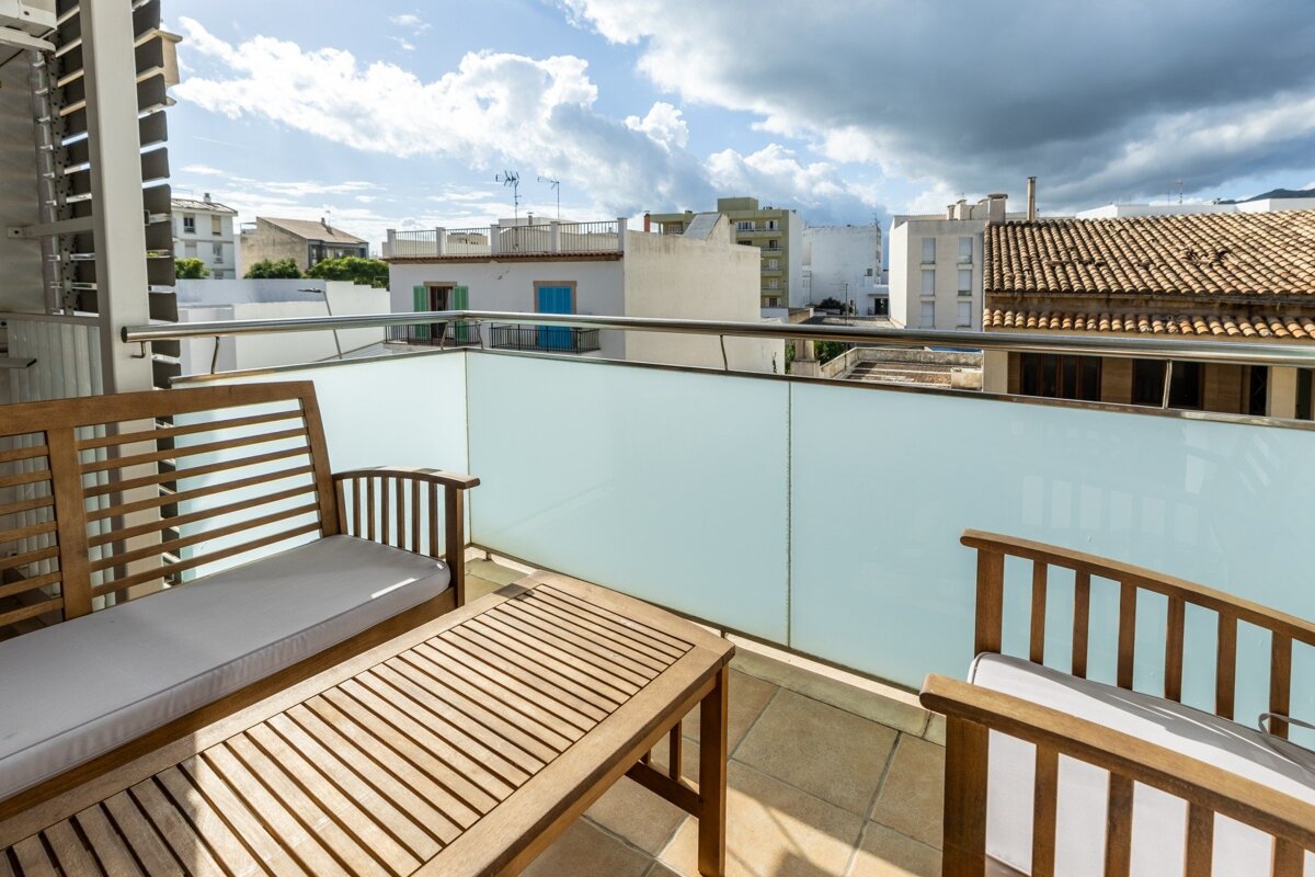 A bright balcony with wooden furniture (bench, chair, coffee table) and a frosted glass railing, overlooking city buildings under a partly cloudy sky.