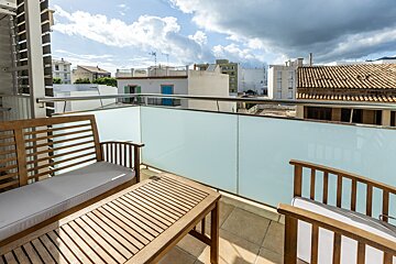 A bright balcony with wooden furniture (bench, chair, coffee table) and a frosted glass railing, overlooking city buildings under a partly cloudy sky.