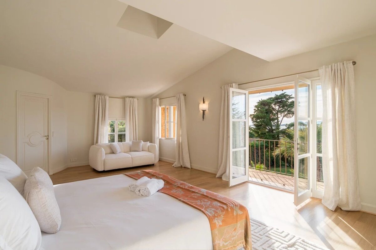 Bright bedroom with a large bed, seating area, and open French doors leading to a balcony overlooking green trees.