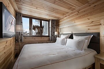 Cozy, rustic wood-paneled bedroom with a large bed, wall-mounted TV showing skiers, and a window overlooking snowy chalets.