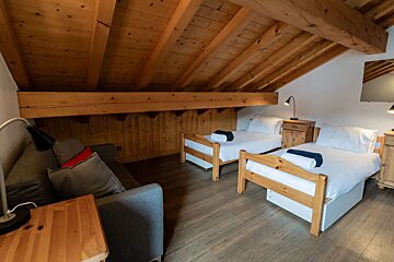 Cozy attic room with exposed wooden beams and light wood flooring. It features two single beds with white linens and a grey sofa with pillows.