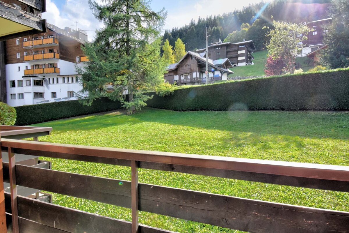Sunny balcony view of a lush green lawn, a long hedge, and chalet-style buildings on a tree-covered mountain slope, with bright sun rays.