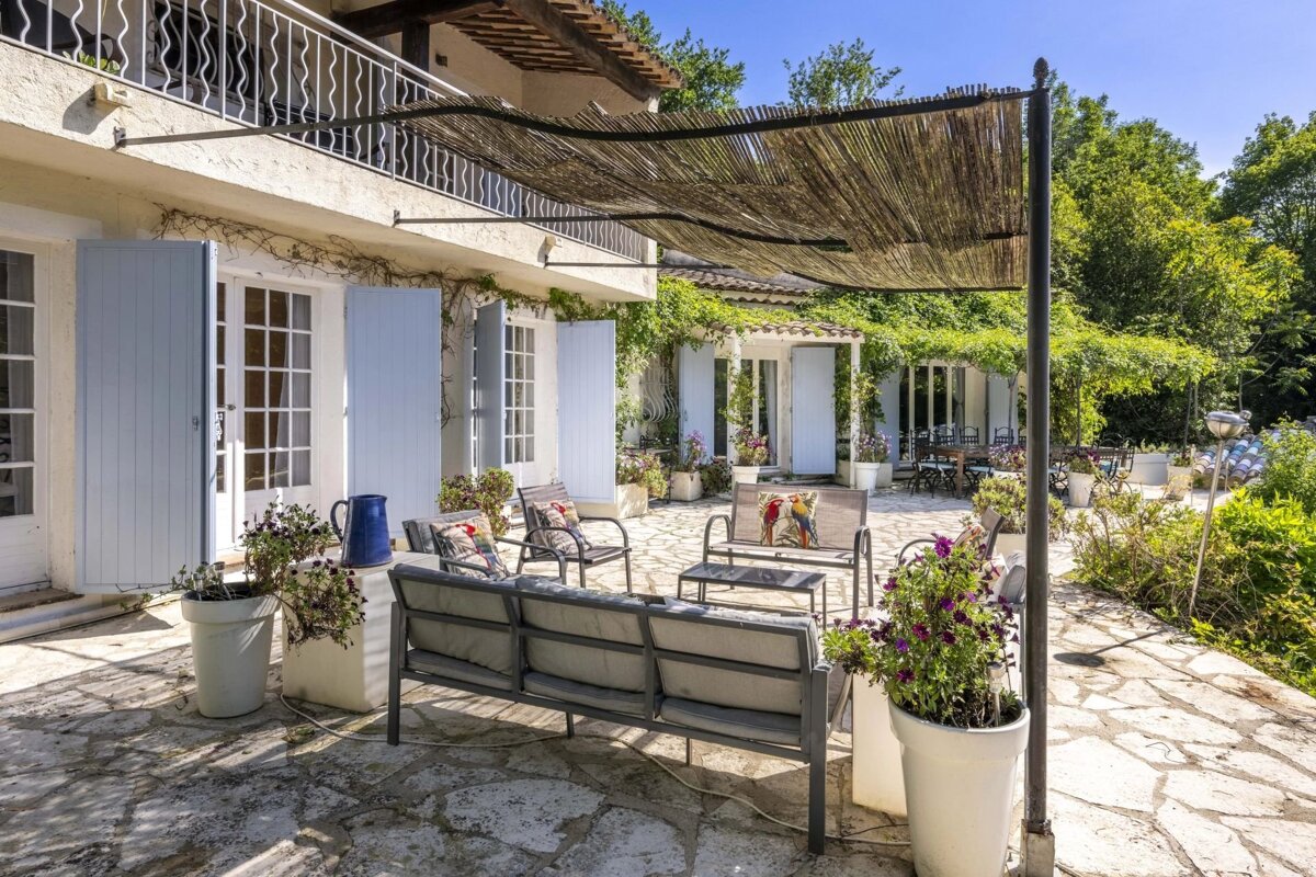 A charming outdoor patio with comfy seating under a bamboo shade, next to a house with blue shutters, surrounded by blooming plants and lush garden.