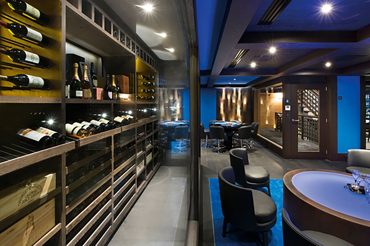 A luxurious modern basement features a well-stocked wine cellar separated by glass from a stylish lounge area with poker tables and blue lighting.
