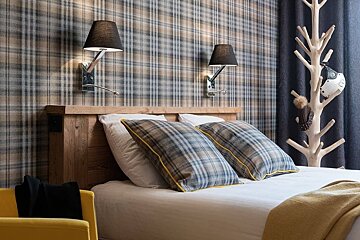 A cozy bedroom with tartan plaid wallpaper and pillows, a rustic wooden headboard, two black lamps, and a tree-shaped coat rack.