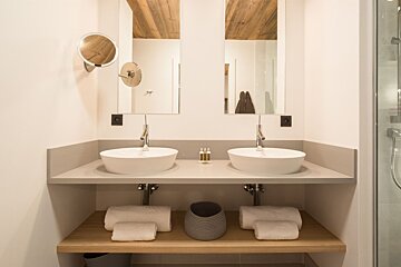 A modern bathroom with dual white vessel sinks, large mirrors, and a wooden ceiling. A light gray counter holds toiletries, with fresh towels below.