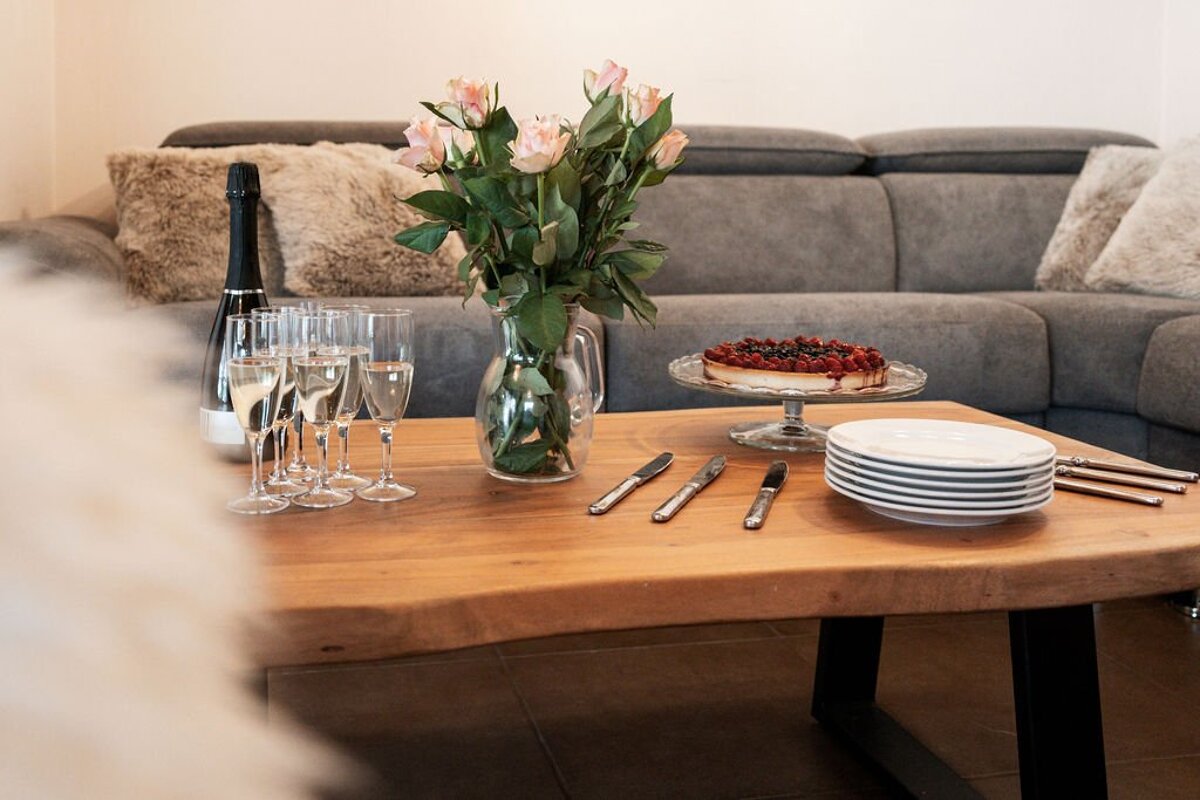 A celebration setup featuring champagne, pink roses, and a berry tart on a wooden coffee table with plates and cutlery, in a cozy living room.