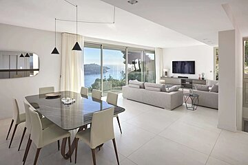 Modern white living/dining room with grey sofas and a dark table, featuring panoramic glass doors overlooking a blue ocean vista.