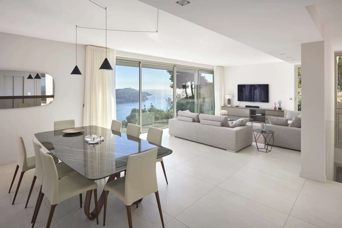 A bright, modern open-plan living and dining room features minimalist grey furniture, a dark dining table, and stunning sea views through large glass doors.