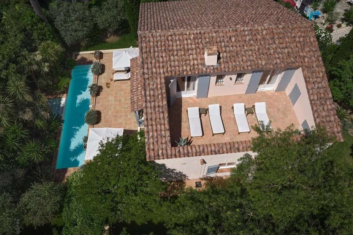 Aerial view of a house with a tiled roof, a large terrace with lounge chairs, and a swimming pool, nestled among green trees.