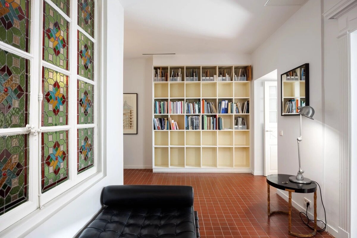 Bright room with a stained-glass window, a black daybed, red tiled floor, and a large grid bookshelf.