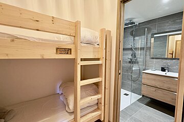 A room with light wooden bunk beds on the left, next to an open doorway revealing a modern bathroom with a shower, sink, and mirror.
