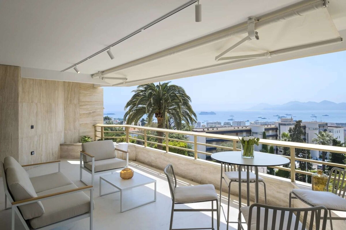 A stylish modern balcony with outdoor furniture, a large palm tree, and a stunning panoramic view of the blue sea and distant city.