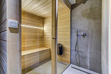 A modern wooden sauna with a glass door stands next to a sleek, grey-tiled shower featuring both rain and handheld showerheads.