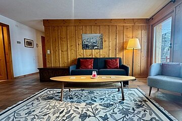 A cozy living room with a wood-paneled wall, blue sofa with red cushions, an oval coffee table, and a floral patterned rug.